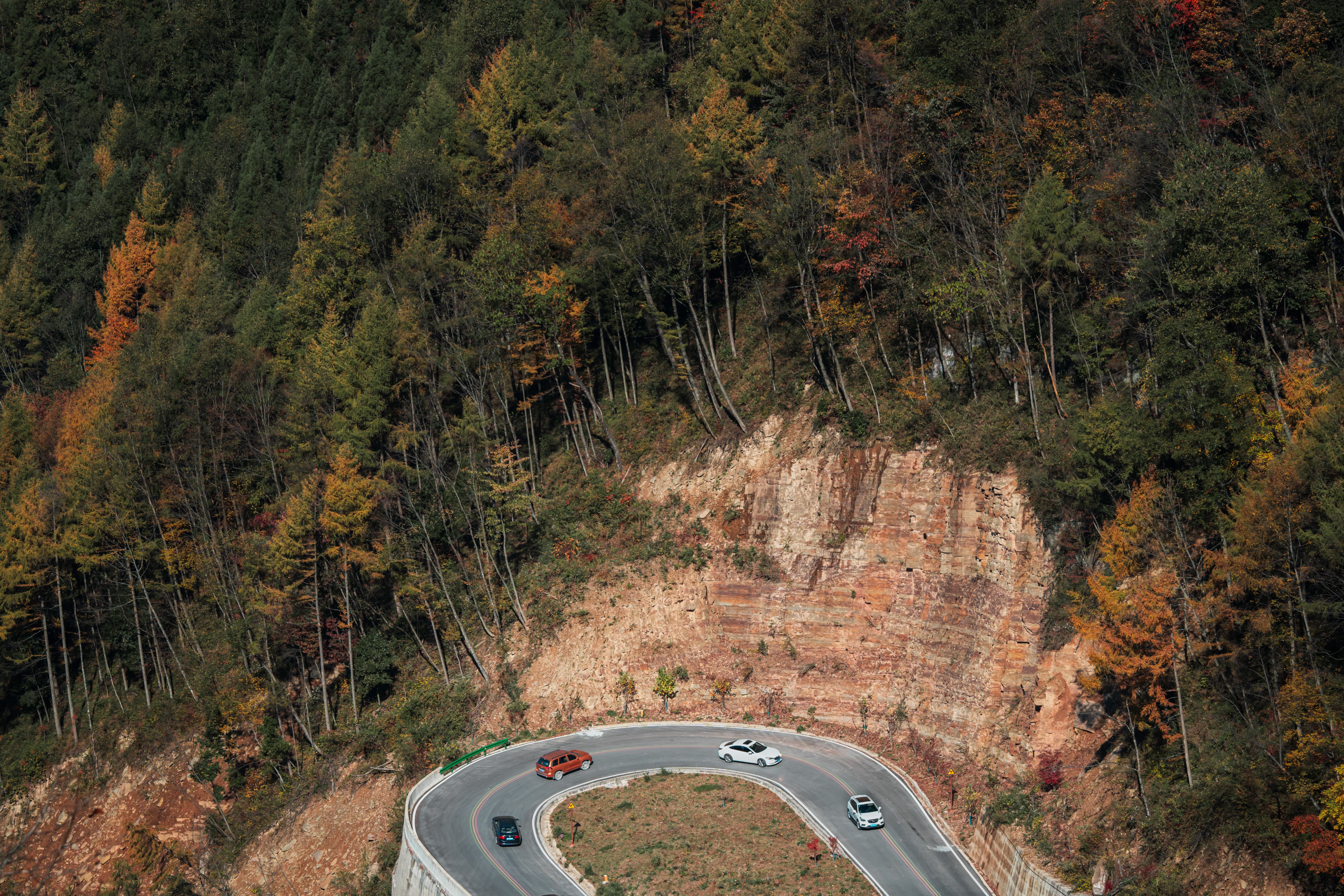 Aerial view of a winding mountain road through autumn trees, with successive bends visible.